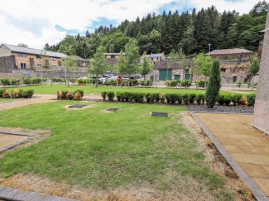 A garden area with grass bushes and trees with houses and forest in the background at Rivers Edge in Newby Bridge