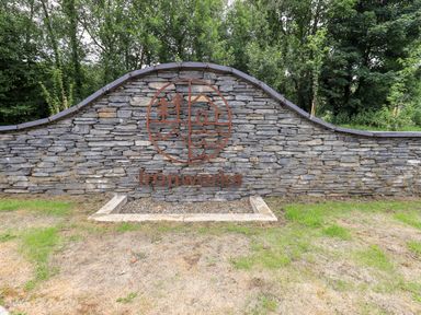 A stone wall with a metal sign displaying trees, waves, a house, and the word Ironworks surrounded by grass and trees at Rivers Edge in Newby Bridge