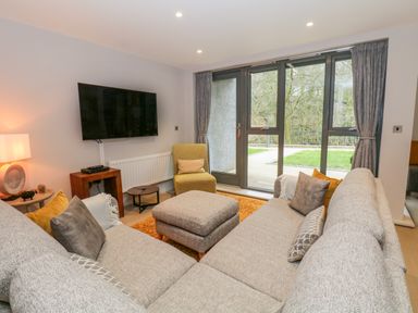 A living room with a large corner sofa and ottoman facing a wall mounted TV and glass doors at Rivers Edge in Newby Bridge