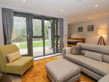 A living room with grey sofa yellow armchair and large windows overlooking a garden at Rivers Edge in Newby Bridge