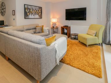 A living room with a gray sectional sofa a yellow armchair a yellow rug a wall-mounted TV and a side table with a lamp at Rivers Edge in Newby Bridge
