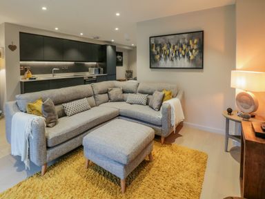 A living room with a grey sectional sofa and ottoman on a yellow rug next to a small round table with a lamp and abstract wall art at Rivers Edge in Newby Bridge
