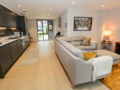 An open plan living room and kitchen with grey sofa and cushions black kitchen cabinets a wall clock and patio doors at Rivers Edge in Newby Bridge