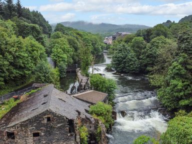 A river flowing past an old stone building surrounded by trees and hills at Rivers Edge in Newby Bridge