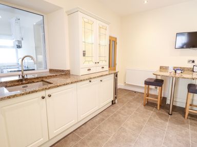A kitchen with a sink a countertop cabinets a glass-fronted cupboard a small table with stools and a wall-mounted television at Craigside in Betws-Y-Coed