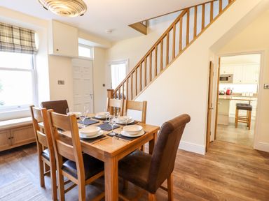 A dining area with a wooden table and six chairs set with plates and glasses near a staircase and an open door to a kitchen at Craigside in Betws-Y-Coed