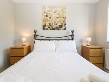 A bedroom with a metal bedframe white bedding wooden bedside tables with lamps and a square mosaic artwork on the wall at 16 Mythern Meadow in Bradford-On-Avon