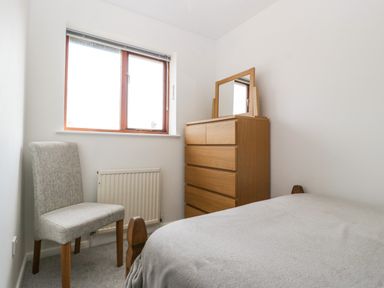 A bedroom with a wooden dresser with a mirror a grey upholstered chair a window and a bed at 16 Mythern Meadow in Bradford-On-Avon