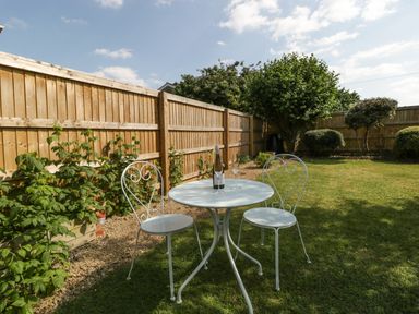 A garden with a white metal table and two chairs on grass with a wooden fence and bushes at 16 Mythern Meadow in Bradford-On-Avon