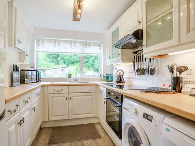 A kitchen with white cabinets, a wooden countertop, a washing machine, oven, microwave, knife set, sink under a window with a floral blind and kitchen utensils at Kavieng Glangrwyney near Gilwern