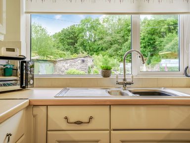 A kitchen sink with a faucet and a microwave on the counter next to a window with a view of trees and a stone wall at Kavieng in Glangrwyney near Gilwern
