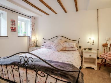 A bedroom with a metal bed frame two nightstands with lamps a chair with towels and a teddy bear and windows with floral curtains at Kavieng in Glangrwyney near Gilwern