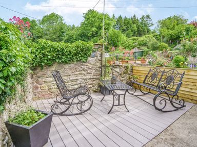 An outdoor seating area with iron rocking chairs and a table on a deck near a stone wall and garden at Kavieng in Glangrwyney near Gilwern