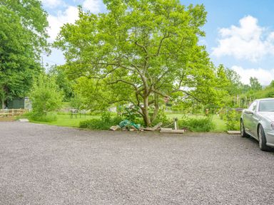 A gravel driveway with a tree in the middle and a silver car parked on the right side with green grass and outdoor seating in the background at Kavieng in Glangrwyney near Gilwern