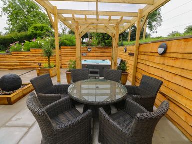 An outdoor patio with six wicker chairs around a glass table under a wooden pergola and a hot tub at Brynawel in Llangynin near Llanboidy
