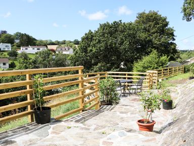An outdoor stone patio with potted plants and metal table and chairs with wooden railing and trees in the background at Rathillet in Feock near Truro