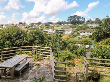 A wooden picnic table and benches on a stone patio with a barbecue and wooden fence overlooking houses trees and boats on water at Rathillet in Feock near Truro