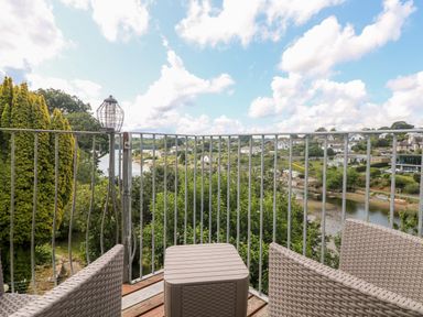 A balcony with woven chairs and a small table overlooking a river and houses at Rathillet in Feock near Truro