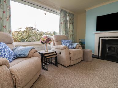 A living room with two beige armchairs a side table with flowers a fireplace and a large window with floral curtains at Rathillet Feock near Truro