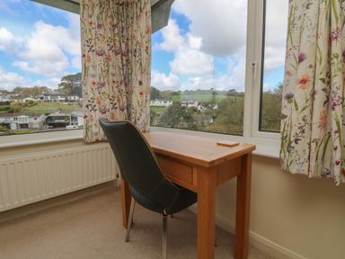 A corner room with a wooden desk and black chair by floral curtains and large windows showing a view of houses and trees at Rathillet in Feock near Truro