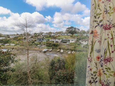 A view of boats on a riverbank with houses and trees under a cloudy sky with floral curtains on the side at Rathillet in Feock near Truro