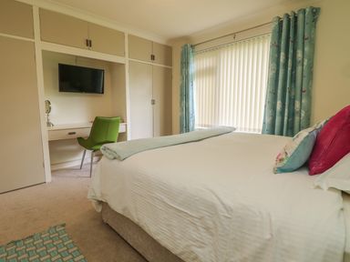 A bedroom with a bed white bedding blue patterned curtains wardrobe a green chair and a wall mounted television at Rathillet in Feock near Truro