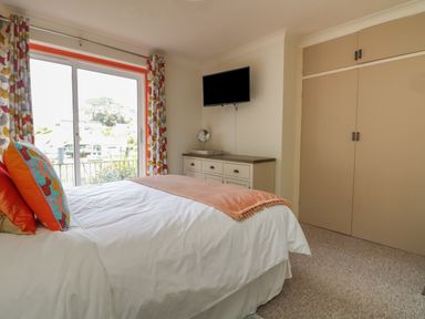 A bedroom with a bed white bedding and orange pillows a dresser with a mirror a wall mounted television and a large window with patterned curtains at Rathillet Feock near Truro