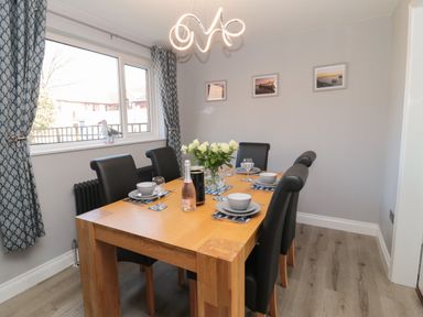 A dining room with a wooden table set for six and a window with blue curtains at Fisherman's Row in Amble