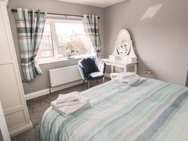 A bedroom with a striped bedcover folded towels a blue chair a white dressing table and striped curtains at Fisherman's Row in Amble