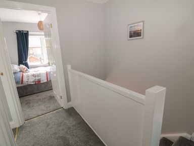 A hallway with carpeted floor and white railing outside a bedroom with a window and dark curtains at Fisherman's Row in Amble