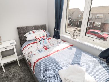 A single bed with patterned bedding and pillows next to a white nightstand and a window with curtains at Fisherman's Row in Amble