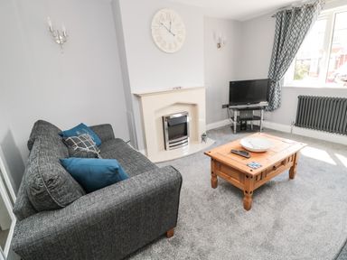 A living room with a grey sofa blue cushions a wooden coffee table a television on a stand a fireplace and a window with patterned curtains at Fisherman's Row in Amble