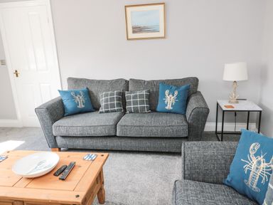 A living room with a gray sofa and blue lobster pillows a wooden coffee table and a side table with a lamp at Fisherman's Row in Amble