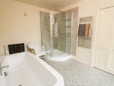 A bathroom with a bathtub a corner shower a white wicker chair and towels on a heated rail at Myrtle Villa in Knighton
