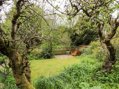 A garden with a wooden swing seat a small table grass and trees at Myrtle Villa in Knighton