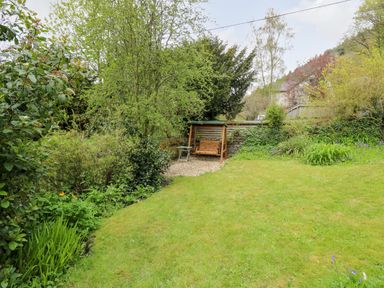 A garden with a wooden swing seat on a gravel area surrounded by trees and bushes at Myrtle Villa in Knighton