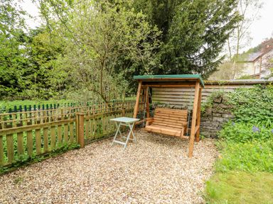 A garden area with a wooden swing bench and a small table on a gravel surface surrounded by trees and fencing at Myrtle Villa in Knighton