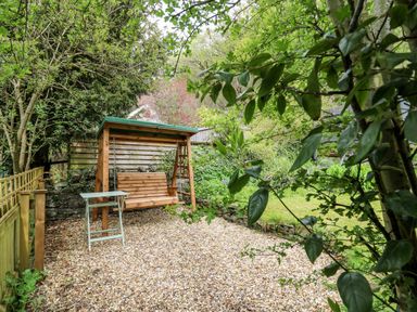 A garden area with a wooden swing bench and small table surrounded by trees at Myrtle Villa in Knighton
