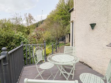 A small outdoor patio with a round metal table and three matching chairs next to a white pebble dash wall with plants and trees in the background at Myrtle Villa in Knighton