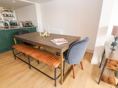 A dining area with a wooden table, two blue chairs, a brown bench, magazines, candle holder, and kitchen counter in the background at 16 Arwenack Street in Falmouth