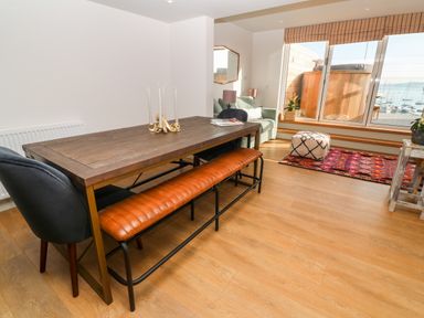 A dining area with a wooden table, leather bench, velvet chair, and a seating area with a sofa, pouf, and large windows overlooking boats at 16 Arwenack Street in Falmouth