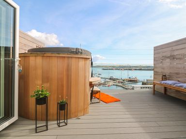 A balcony with a wooden hot tub two potted plants a wooden bench with cushions a small table with an orange rug and a view of boats on the water at 16 Arwenack Street in Falmouth