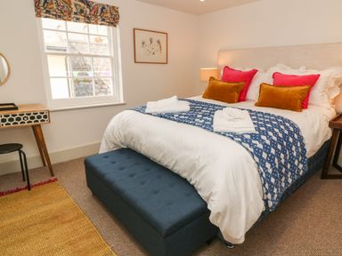 A bedroom with a bed with colorful pillows a blue bench at the foot of the bed a window with a patterned valance and a small desk at 16 Arwenack Street in Falmouth
