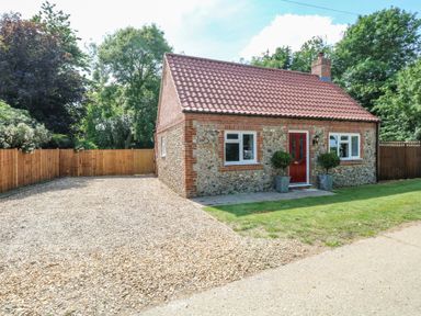 A small brick house with a red door and gravel driveway surrounded by wooden fences and trees at Keepers Cottage Beeston near Dereham