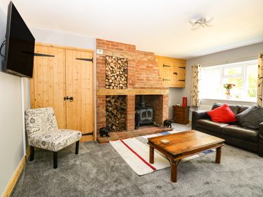 A living room with a brick fireplace stacked with firewood a wooden coffee table a leather sofa a patterned chair and a wall-mounted TV at Keepers Cottage in Beeston near Dereham