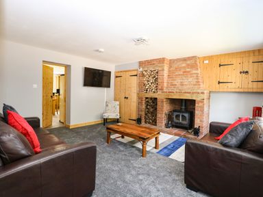 A living room with brown leather sofas red and gray pillows a wooden coffee table a brick fireplace and a wall mounted TV at Keepers Cottage in Beeston near Dereham