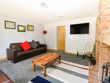 A living room with a brown sofa with red and gray cushions a wooden coffee table a patterned chair a wall mounted television and a brick fireplace at Keepers Cottage Beeston near Dereham