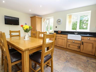 A kitchen with a wooden dining table and chairs a vase with flowers on the table wooden cabinets a sink under the window and a wall mounted television at Keepers Cottage Beeston near Dereham