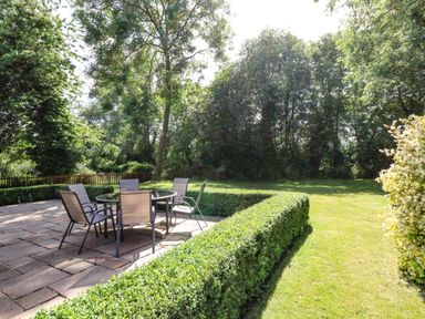 An outdoor patio area with a table and six chairs surrounded by hedges and a grassy lawn at Keepers Cottage Beeston near Dereham