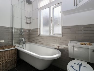 A bathroom with a bathtub a wicker laundry basket a window and a toilet with an anchor design on the seat at Ferriwais in Appledore